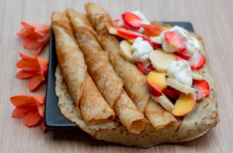 Three rolled crepes are placed on a square black plate with an additional unfolded crepe beside them. The unfolded crepe is topped with slices of strawberries, bananas, and peaches, along with dollops of whipped cream. Bright orange flowers artfully decorate the backdrop on the wooden table surface.