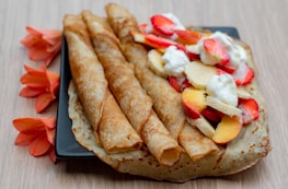 Three rolled crepes are placed on a square black plate with an additional unfolded crepe beside them. The unfolded crepe is topped with slices of strawberries, bananas, and peaches, along with dollops of whipped cream. Bright orange flowers artfully decorate the backdrop on the wooden table surface.