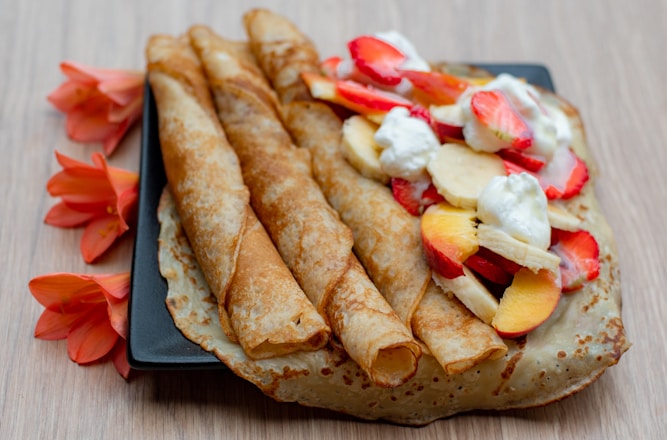 Three rolled crepes are placed on a square black plate with an additional unfolded crepe beside them. The unfolded crepe is topped with slices of strawberries, bananas, and peaches, along with dollops of whipped cream. Bright orange flowers artfully decorate the backdrop on the wooden table surface.