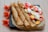 Three rolled crepes are placed on a square black plate with an additional unfolded crepe beside them. The unfolded crepe is topped with slices of strawberries, bananas, and peaches, along with dollops of whipped cream. Bright orange flowers artfully decorate the backdrop on the wooden table surface.
