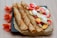 Three rolled crepes are placed on a square black plate with an additional unfolded crepe beside them. The unfolded crepe is topped with slices of strawberries, bananas, and peaches, along with dollops of whipped cream. Bright orange flowers artfully decorate the backdrop on the wooden table surface.