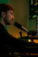 A person is sitting in front of a computer screen, speaking into a microphone. The environment is dimly lit with warm, yellow and green hues, creating a focused atmosphere. Audio equipment and the edge of a computer monitor are visible in the background.