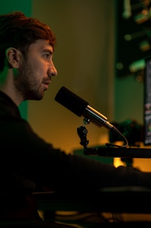 A person is sitting in front of a computer screen, speaking into a microphone. The environment is dimly lit with warm, yellow and green hues, creating a focused atmosphere. Audio equipment and the edge of a computer monitor are visible in the background.