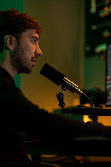 A person is sitting in front of a computer screen, speaking into a microphone. The environment is dimly lit with warm, yellow and green hues, creating a focused atmosphere. Audio equipment and the edge of a computer monitor are visible in the background.