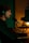 A person wearing headphones sits at a desk typing on a keyboard, in front of a large microphone. There is a camera setup in the background next to a monitor displaying text, and the scene is dimly lit with a warm and greenish ambiance.