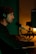 A person wearing headphones sits at a desk typing on a keyboard, in front of a large microphone. There is a camera setup in the background next to a monitor displaying text, and the scene is dimly lit with a warm and greenish ambiance.