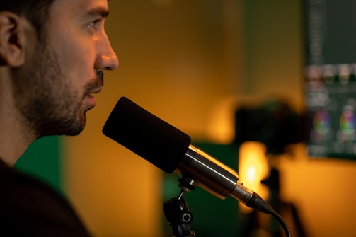 A person is speaking into a microphone, with a blurred background suggesting the setting is a recording or broadcast studio. The lighting is warm, creating a focused and professional atmosphere. A computer screen with editing software is partially visible in the background.