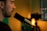 A person is speaking into a microphone, with a blurred background suggesting the setting is a recording or broadcast studio. The lighting is warm, creating a focused and professional atmosphere. A computer screen with editing software is partially visible in the background.