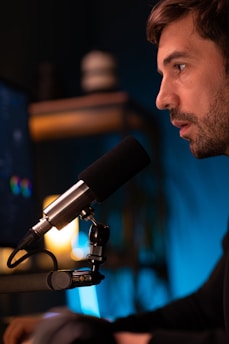 A person sitting in front of a microphone, appearing focused and engaged. The background includes a blurred shelf with warm lighting, adding to the intimate atmosphere.