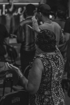 A group of people is gathered, with one person in the foreground raising their hands, possibly in prayer or contemplation. The setting appears to be indoors with seats placed closely together.
