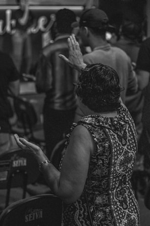 A group of people is gathered, with one person in the foreground raising their hands, possibly in prayer or contemplation. The setting appears to be indoors with seats placed closely together.