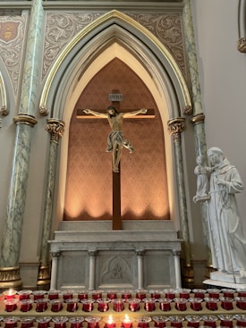An ornate altar with a crucifix prominently displayed. The crucifix is set against a decorative pattern with gold accents, surrounded by marble columns. Below the crucifix, there is a collection of red votive candles on a stand. To the right, a white statue of a religious figure holding a child is visible.