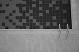 A tiled swimming pool is partly filled with water, creating a pattern of dark and light squares on the surface. A metal pool ladder is affixed to the side, and the surrounding area is paved with light-colored tiles.