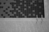 A tiled swimming pool is partly filled with water, creating a pattern of dark and light squares on the surface. A metal pool ladder is affixed to the side, and the surrounding area is paved with light-colored tiles.