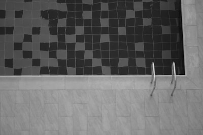 A tiled swimming pool is partly filled with water, creating a pattern of dark and light squares on the surface. A metal pool ladder is affixed to the side, and the surrounding area is paved with light-colored tiles.
