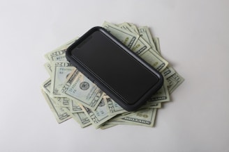 A smartphone is placed on top of a pile of US twenty dollar bills, creating a contrast between digital technology and physical currency. The bills are scattered and visible from various angles.
