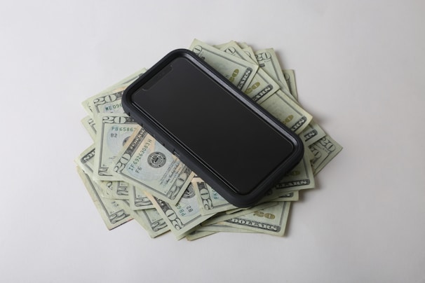 A smartphone is placed on top of a pile of US twenty dollar bills, creating a contrast between digital technology and physical currency. The bills are scattered and visible from various angles.