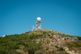Situated atop a hill, a large radar tower featuring a dome-shaped top is surrounded by multiple antennas and satellite dishes. The foreground consists of rocky terrain with patches of greenery under a clear blue sky.