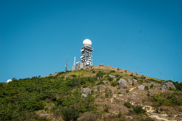 Situated atop a hill, a large radar tower featuring a dome-shaped top is surrounded by multiple antennas and satellite dishes. The foreground consists of rocky terrain with patches of greenery under a clear blue sky.