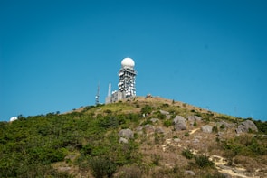 Situated atop a hill, a large radar tower featuring a dome-shaped top is surrounded by multiple antennas and satellite dishes. The foreground consists of rocky terrain with patches of greenery under a clear blue sky.
