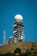 A large radar tower stands prominently on a hilltop, featuring a spherical dome at the top and multiple satellite dishes attached to its metal structure. The base of the tower is surrounded by a rocky, grassy terrain with scattered shrubs and bushes. The sky is clear and blue, providing a sharp contrast to the white and metallic elements of the tower.