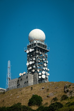 A large radar tower stands prominently on a hilltop, featuring a spherical dome at the top and multiple satellite dishes attached to its metal structure. The base of the tower is surrounded by a rocky, grassy terrain with scattered shrubs and bushes. The sky is clear and blue, providing a sharp contrast to the white and metallic elements of the tower.