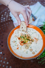 A hand holds a piece of bread over a bowl filled with creamy hummus garnished with chickpeas, chopped herbs, and a sprinkle of paprika. The orange rim of the bowl complements the rustic table surface. A green plant and a folded napkin are visible in the background.