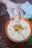 A hand holds a piece of bread over a bowl filled with creamy hummus garnished with chickpeas, chopped herbs, and a sprinkle of paprika. The orange rim of the bowl complements the rustic table surface. A green plant and a folded napkin are visible in the background.