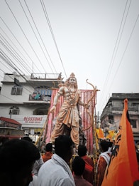 A large statue of a deity stands amidst a crowd in a street setting. The statue is adorned with garlands and appears to be on a decorated platform. Several people, some dressed in traditional attire, gather around. Orange flags with black symbols are prominently displayed, blending with the festive and religious atmosphere. Buildings and power lines form the backdrop.