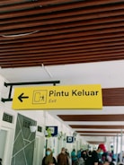 A yellow sign with the words 'Pintu Keluar' and 'Exit' is shown, featuring an arrow pointing to the left and a pictogram representing an exit. Below the sign, people are seen walking and wearing face masks. The ceiling has brown wooden beams, and the walls have decorative elements and hanging planters.
