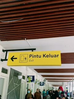A yellow sign with the words 'Pintu Keluar' and 'Exit' is shown, featuring an arrow pointing to the left and a pictogram representing an exit. Below the sign, people are seen walking and wearing face masks. The ceiling has brown wooden beams, and the walls have decorative elements and hanging planters.