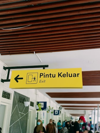 A yellow sign with the words 'Pintu Keluar' and 'Exit' is shown, featuring an arrow pointing to the left and a pictogram representing an exit. Below the sign, people are seen walking and wearing face masks. The ceiling has brown wooden beams, and the walls have decorative elements and hanging planters.