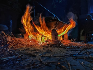 Bright orange and yellow flames burn under a makeshift stove setup, with dry twigs and sticks scattered around. The fire illuminates the surroundings, highlighting the dark, rustic atmosphere.
