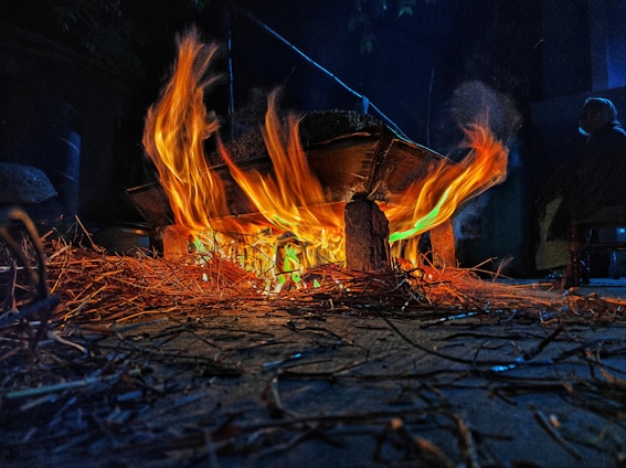 Bright orange and yellow flames burn under a makeshift stove setup, with dry twigs and sticks scattered around. The fire illuminates the surroundings, highlighting the dark, rustic atmosphere.