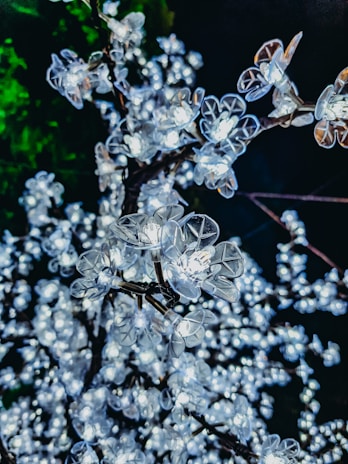A close-up view of artificial flowers with built-in lights, resembling cherry blossoms on branches. The flowers are illuminated, creating a bright and sparkling effect against a dark background with a hint of green foliage.