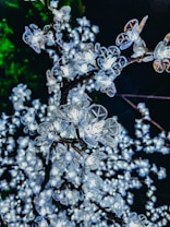 A close-up view of artificial flowers with built-in lights, resembling cherry blossoms on branches. The flowers are illuminated, creating a bright and sparkling effect against a dark background with a hint of green foliage.