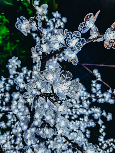 A close-up view of artificial flowers with built-in lights, resembling cherry blossoms on branches. The flowers are illuminated, creating a bright and sparkling effect against a dark background with a hint of green foliage.