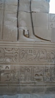 The image displays a section of an ancient stone wall adorned with intricate Egyptian hieroglyphs and carvings. The carvings include symbols, birds, and figures, showcasing traditional Egyptian artistry. A large carved leg and foot are also present as part of a larger sculpture.