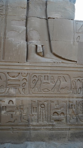 The image displays a section of an ancient stone wall adorned with intricate Egyptian hieroglyphs and carvings. The carvings include symbols, birds, and figures, showcasing traditional Egyptian artistry. A large carved leg and foot are also present as part of a larger sculpture.