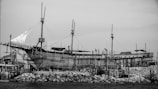 An old wooden ship is under construction on a rocky shore. The ship features multiple masts and a sail partially unfurled, surrounded by scaffolding and structures that support the shipbuilding process. The scene conveys a sense of craftsmanship and maritime tradition.