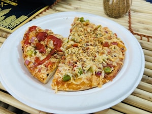 A small pizza with a variety of toppings including cheese, green peppers, onions, and bits of red vegetables, appears on a white paper plate. One half is drizzled with a red sauce, possibly ketchup. The pizza is set on a bamboo mat, with a black and gold menu in the background and a decorative vase nearby.