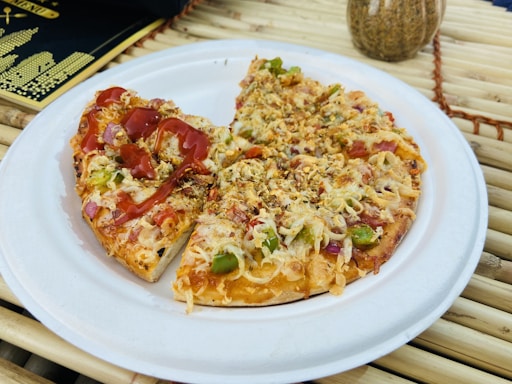 A small pizza with a variety of toppings including cheese, green peppers, onions, and bits of red vegetables, appears on a white paper plate. One half is drizzled with a red sauce, possibly ketchup. The pizza is set on a bamboo mat, with a black and gold menu in the background and a decorative vase nearby.