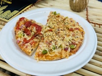 A small pizza with a variety of toppings including cheese, green peppers, onions, and bits of red vegetables, appears on a white paper plate. One half is drizzled with a red sauce, possibly ketchup. The pizza is set on a bamboo mat, with a black and gold menu in the background and a decorative vase nearby.