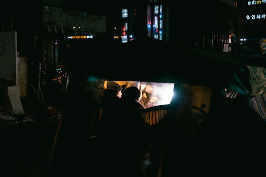 A dimly lit urban scene at night with people inside a small tent or shelter. The inside of the tent is illuminated, creating a stark contrast with the dark surroundings. Neon signs and lights from nearby buildings are visible in the background, adding a sense of urban life.