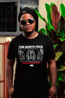 A person wearing sunglasses and a black beanie stands outdoors in front of a window and large green leaves. The individual is wearing a black t-shirt with white and red text and graphic designs featuring jackets. The setting suggests a casual environment with plant life.