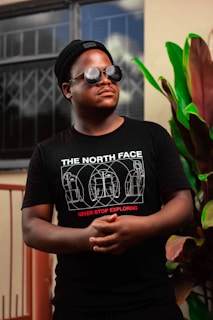 A person wearing a black beanie, reflective sunglasses, and a black T-shirt with a graphic featuring backpacks and the slogan 'Never Stop Exploring'. The background includes a window with a grille and a large plant with green and burgundy leaves.