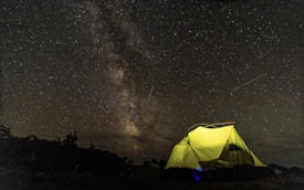A brightly lit tent is set up under a vast, starry night sky with a prominent view of the Milky Way galaxy. The tent's yellow illumination contrasts with the dark silhouette of surrounding vegetation and landscape. Shooting stars or meteors are visible across the sky.