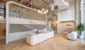 A modern reception area with a sleek, curved reception desk at the center, decorated with wicker baskets and a vase containing small decorative objects. The background features an arched structure with vertical wooden slats that house the sign 'the bureau.' To the right, a cozy seating area includes two white armchairs, a small round table, and potted plants. The ceiling is adorned with industrial-style exposed pipes and ducts, with hanging lights providing illumination. The floor is designed with a herringbone pattern.