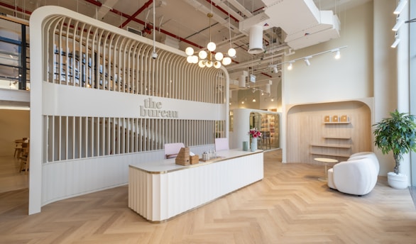 A modern reception area with a sleek, curved reception desk at the center, decorated with wicker baskets and a vase containing small decorative objects. The background features an arched structure with vertical wooden slats that house the sign 'the bureau.' To the right, a cozy seating area includes two white armchairs, a small round table, and potted plants. The ceiling is adorned with industrial-style exposed pipes and ducts, with hanging lights providing illumination. The floor is designed with a herringbone pattern.