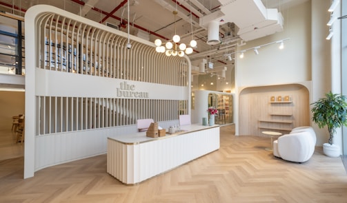 A modern reception area with a sleek, curved reception desk at the center, decorated with wicker baskets and a vase containing small decorative objects. The background features an arched structure with vertical wooden slats that house the sign 'the bureau.' To the right, a cozy seating area includes two white armchairs, a small round table, and potted plants. The ceiling is adorned with industrial-style exposed pipes and ducts, with hanging lights providing illumination. The floor is designed with a herringbone pattern.